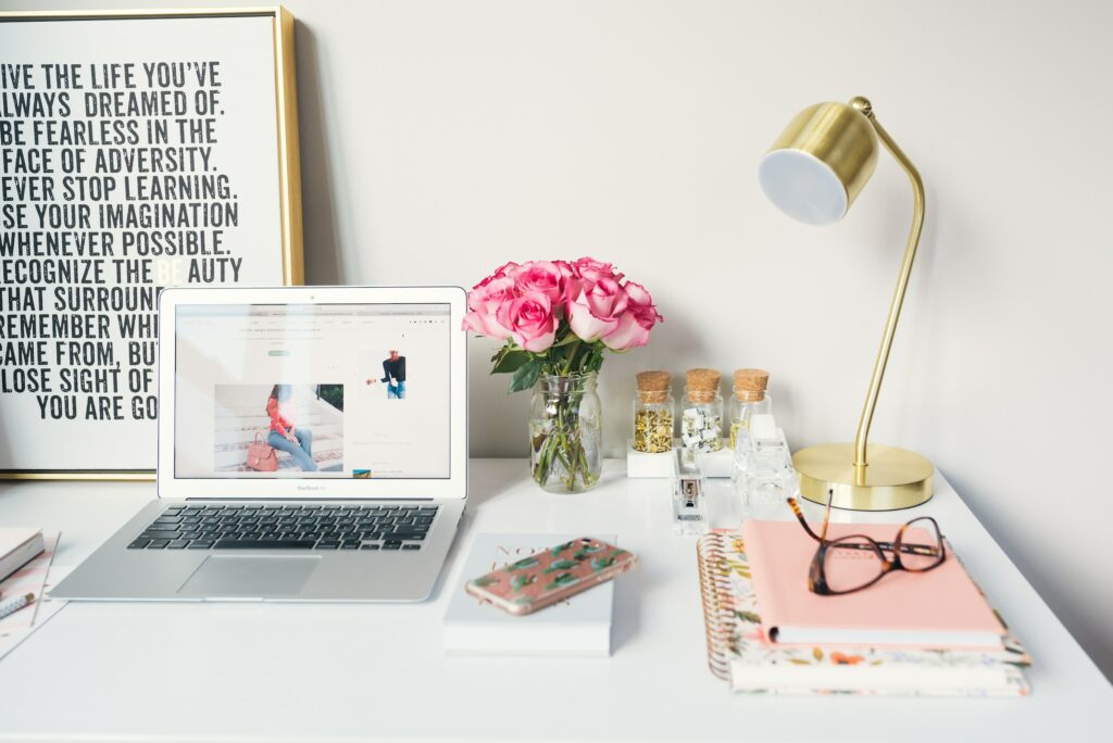 work desk showing a laptop, lamp, flowers and notebooks